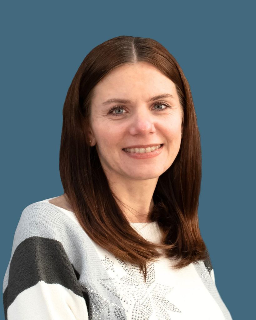 A woman with straight brown hair smiles slightly, wearing a light sweater with dark stripes on a blue background.
