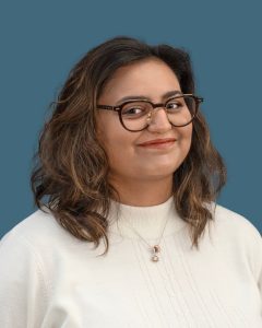 Person with shoulder-length hair and glasses, wearing a white sweater and a necklace, smiles slightly against a solid blue background. Person with shoulder-length hair and glasses, wearing a white sweater and a necklace, smiles slightly against a solid blue background.