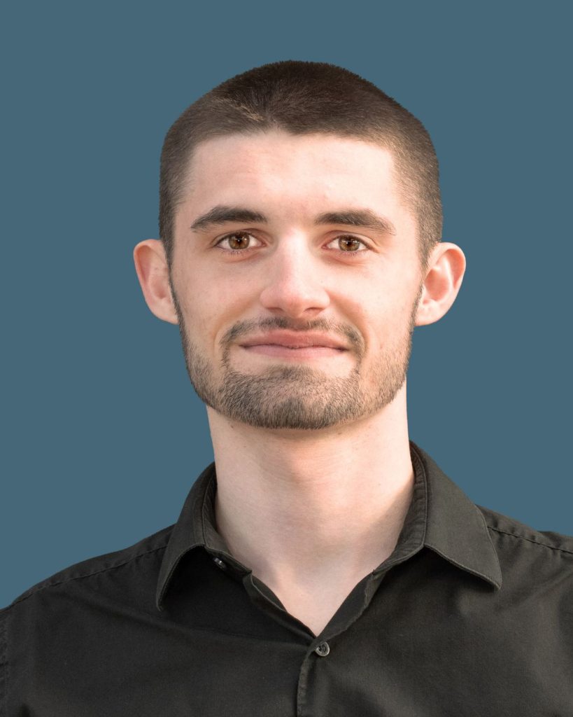 A man with short hair and beard wearing a black collared shirt against a plain blue background.