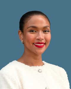 Person with short hair wearing a cream-colored jacket against a blue background, smiling. Person with short hair wearing a cream-colored jacket against a blue background, smiling.